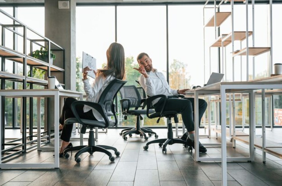 a young woman and man sitting in office chairs, chatting, and smiling in a modern office