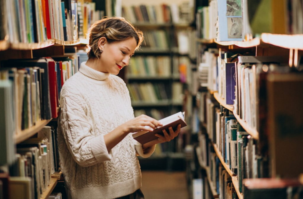 A smiling woman in a cozy sweater browses a book in a library aisle