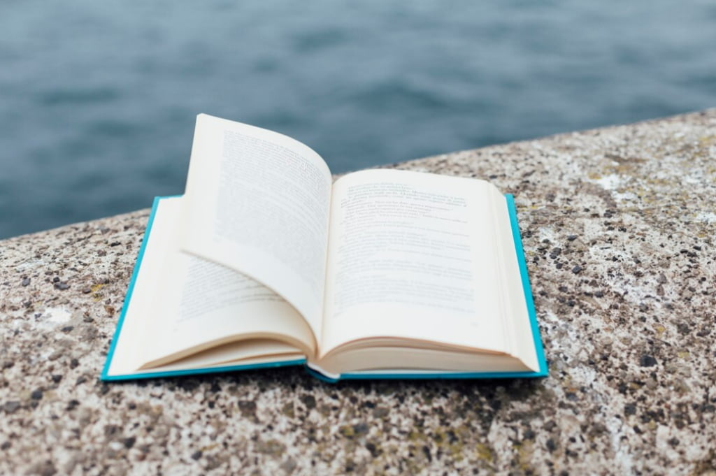 An open book lies on a concrete surface against a backdrop of water