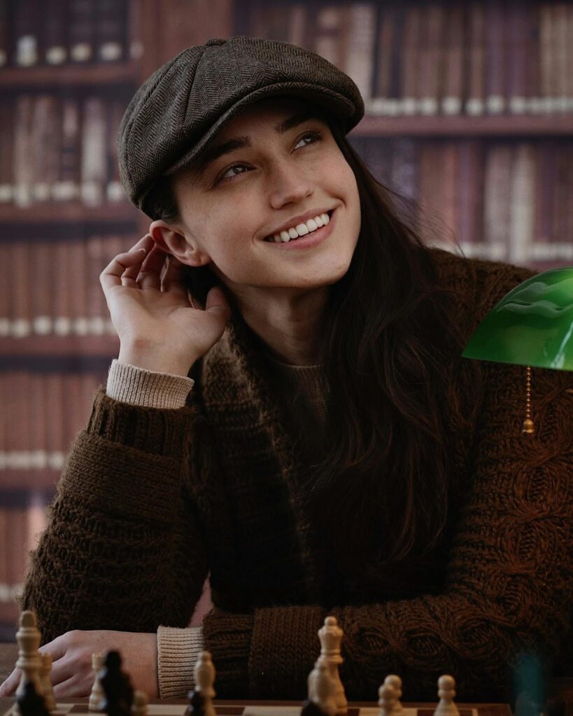 A girl in a sweater and a cape sits against the background of a bookcase, with a chessboard in front of her