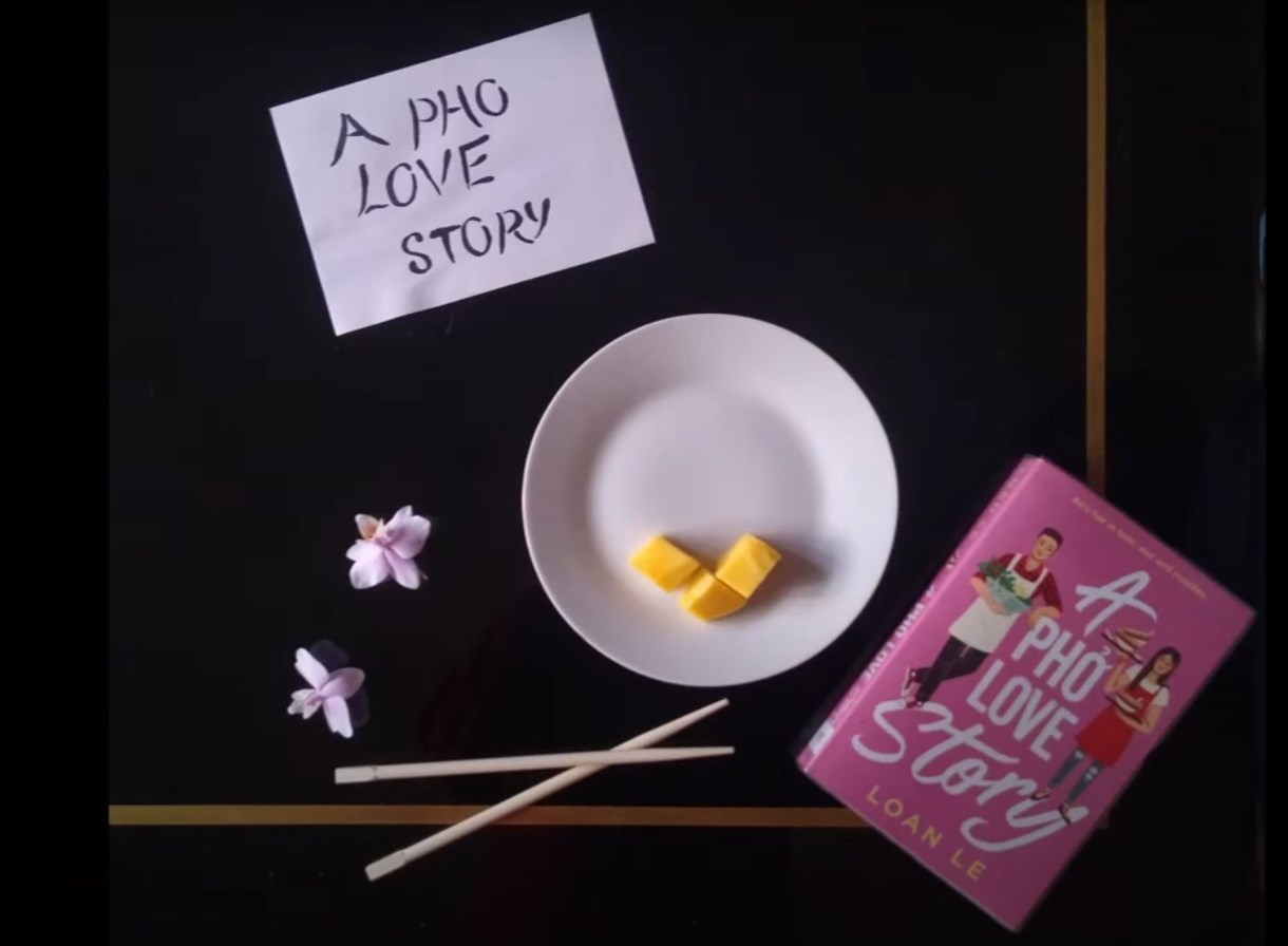 ‘A Pho Love Story” book by Loan Le, with a white plate, cheese pieces, chopsticks, and flowers on a black background
